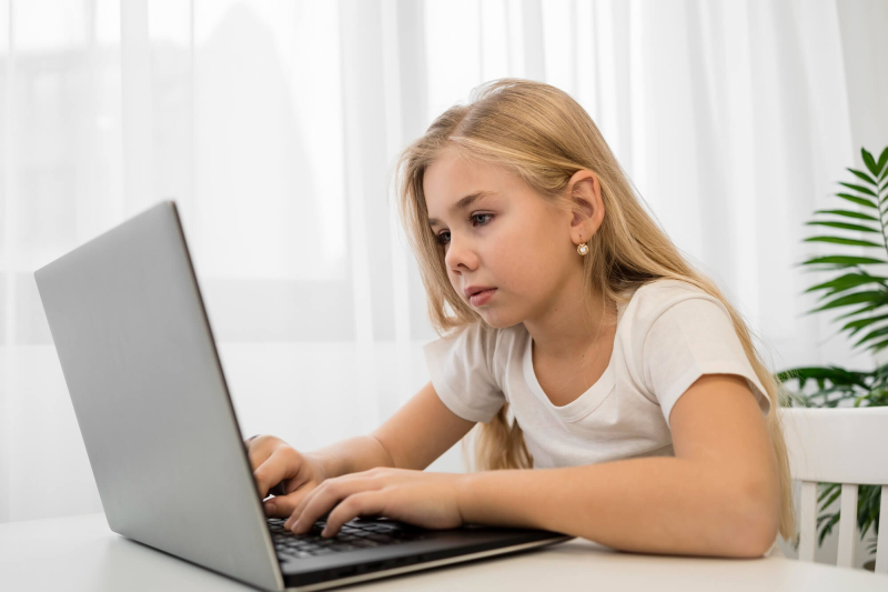 Dziewczynka w wieku szkolnym siedzi przy biurku i pisze na laptopie.
