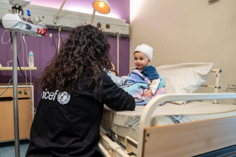 Pracownica UNICEF kuca przy łóżku szpitalnym obok 10-miesięcznego Ahmada, który odniósł obrażenia podczas izraelskiego nalotu.