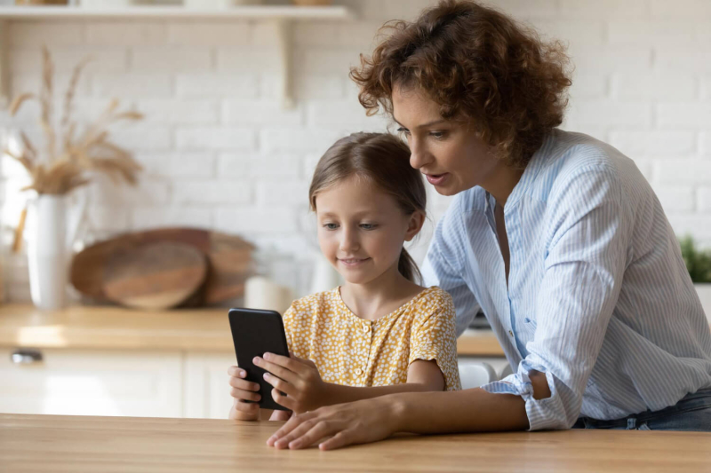 Mama i c&oacute;rka patrzą w ekran trzymanego przez dziewczynkę smartfona.