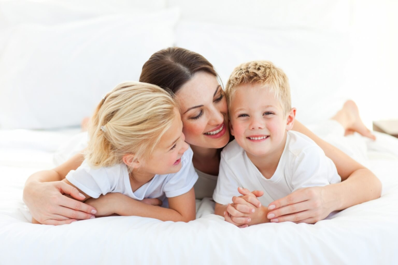 Mama przytula uśmiechnięte dzieci podczas odpoczynku na hotelowym ł&oacute;żku.