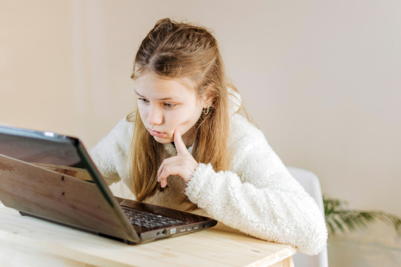 Dziewczynka w wieku szkolnym siedzi przy biurku, patrząc w ekran laptopa.