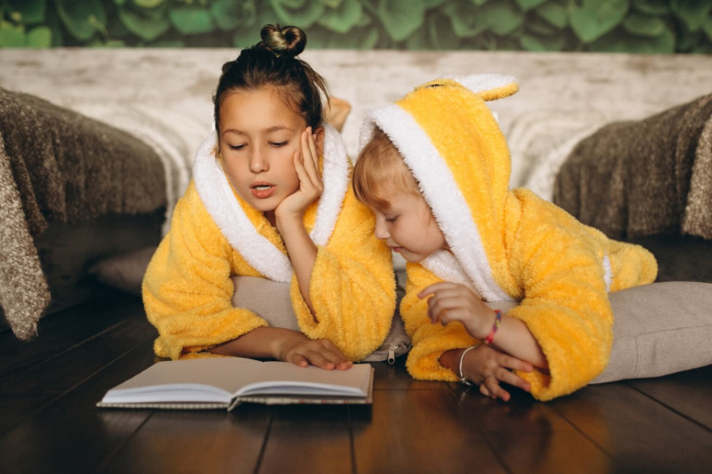 Dwoje dzieci odpoczywa w hotelu, wsp&oacute;lnie czytając książkę.