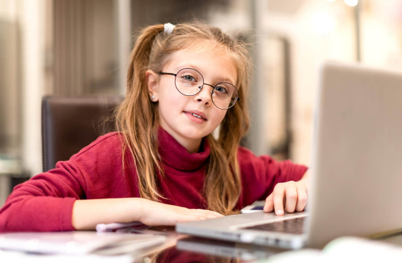 Dziewczynka w wielu szkolnym siedzi przy biurku i odrabia lekcje przy użyciu laptopa.